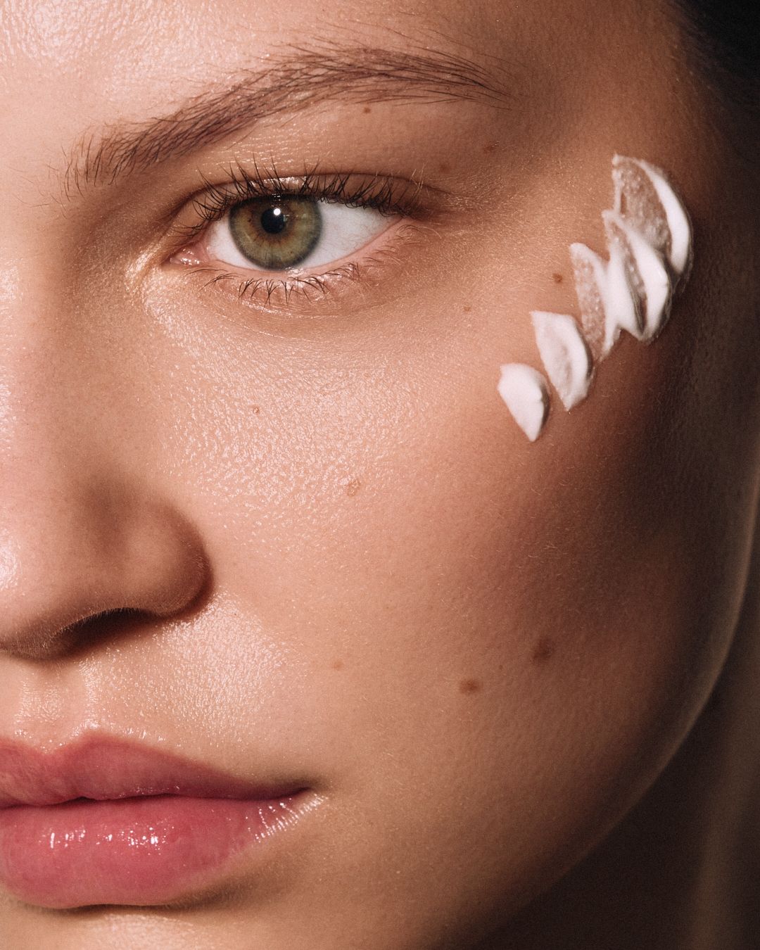 Close-up of a woman's face with green eyes and white cream swatches applied to her cheekbone for skincare.