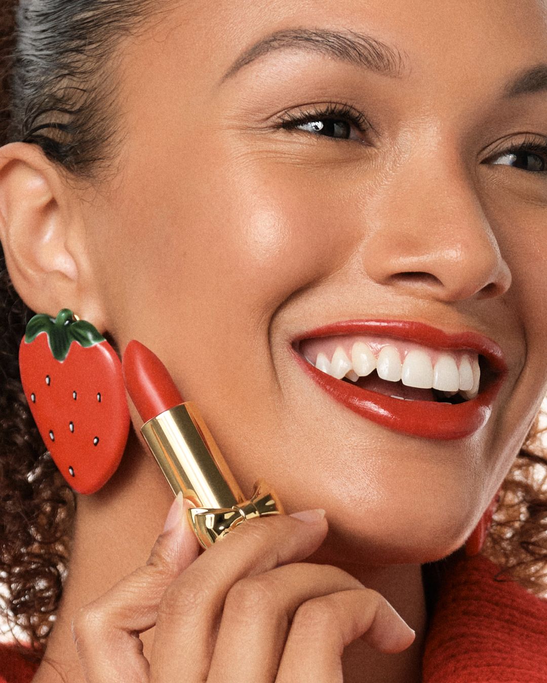 A joyful woman holding a red lipstick near her face, wearing a strawberry earring, showcasing a bold red lip.