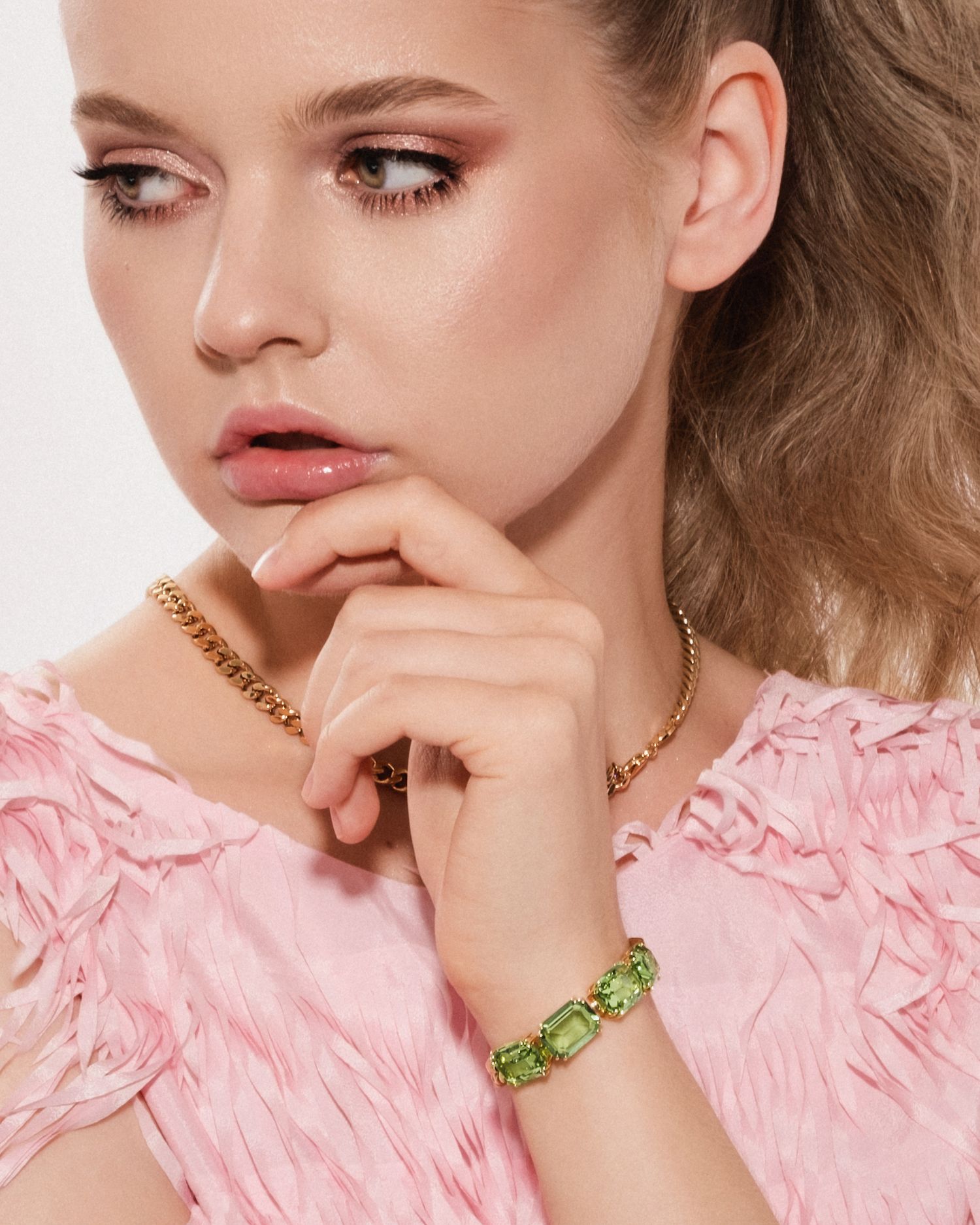 Model wearing gold chain necklace and green crystal bracelet with pink ruffled top, posing thoughtfully.
