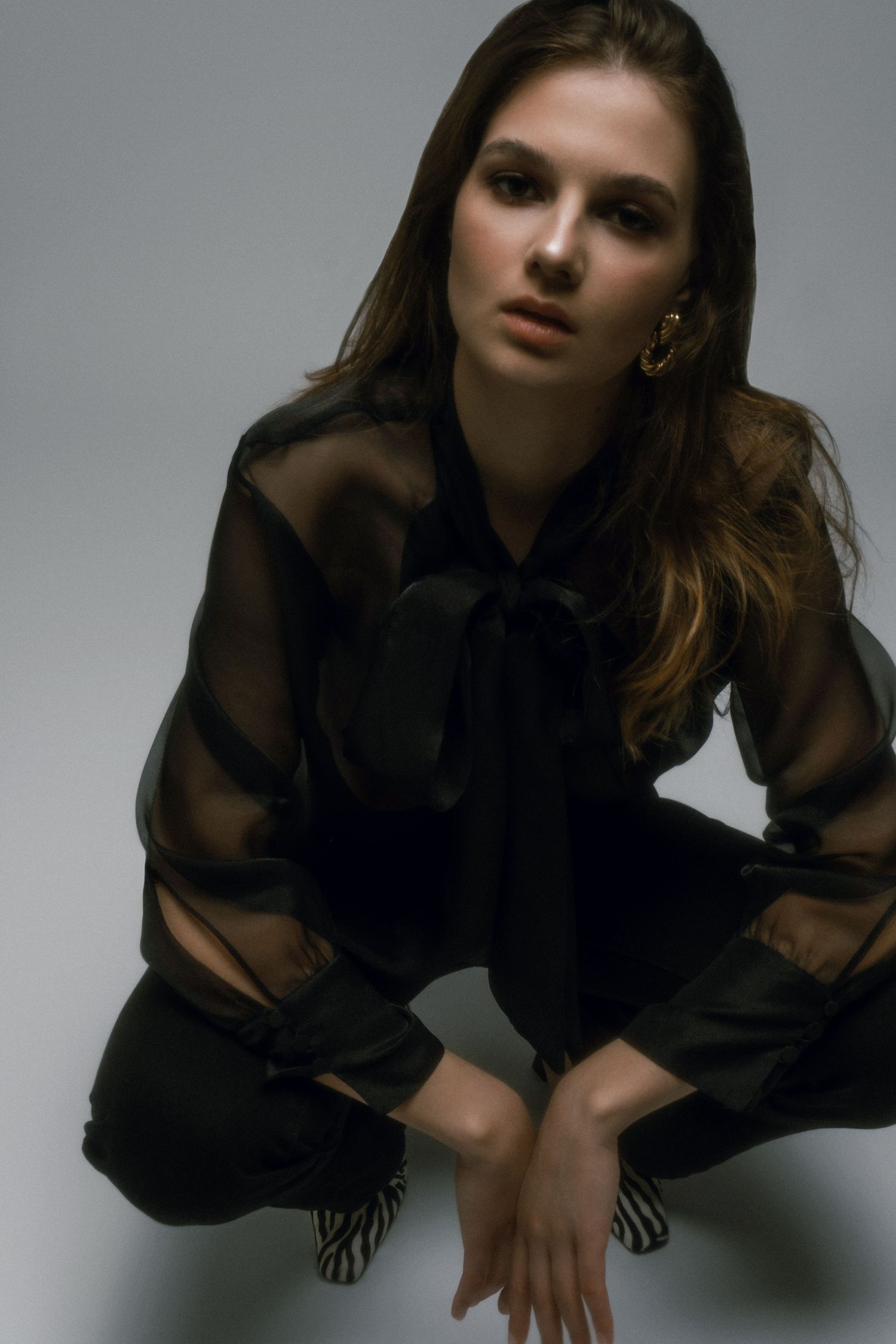 Young woman in sheer black blouse with bow tie and black pants sitting gracefully on studio floor.