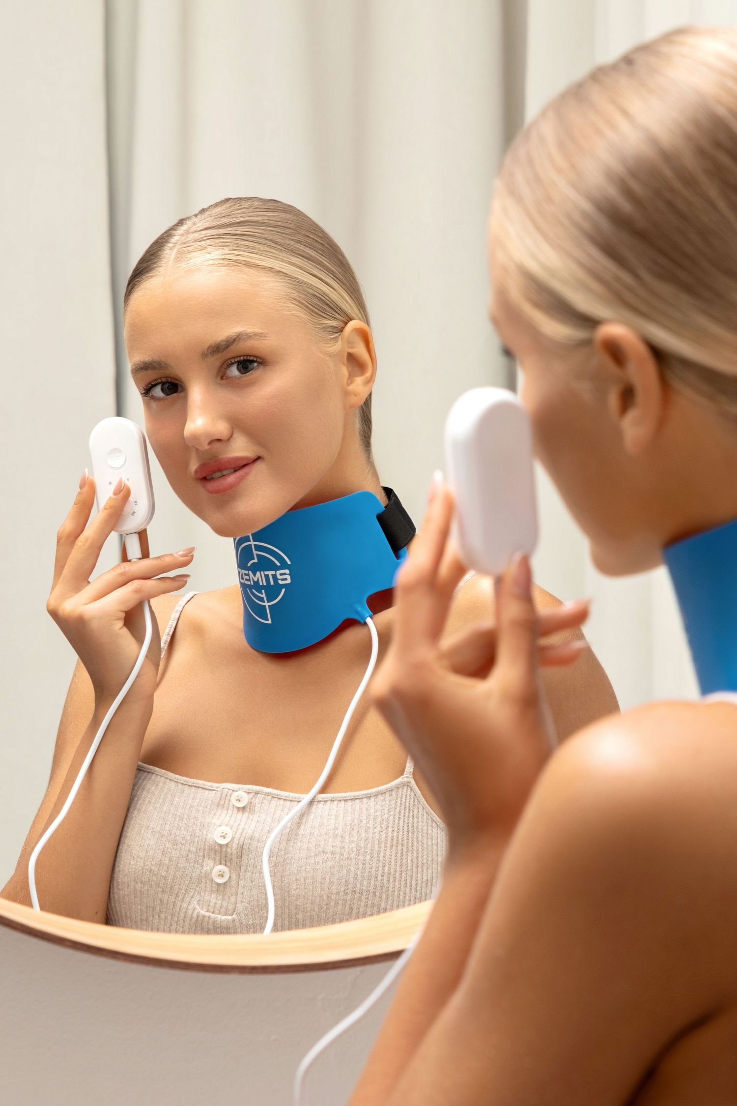Young woman wearing a blue neck massager device, holding a controller while looking in a mirror.