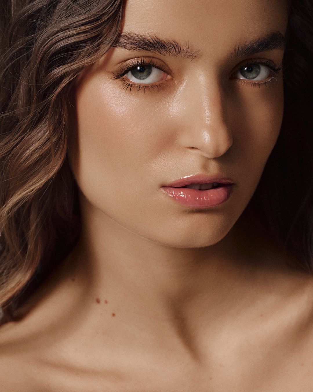 A close-up portrait of a woman with wavy brown hair, blue-gray eyes, glowing skin, and glossy nude lips.