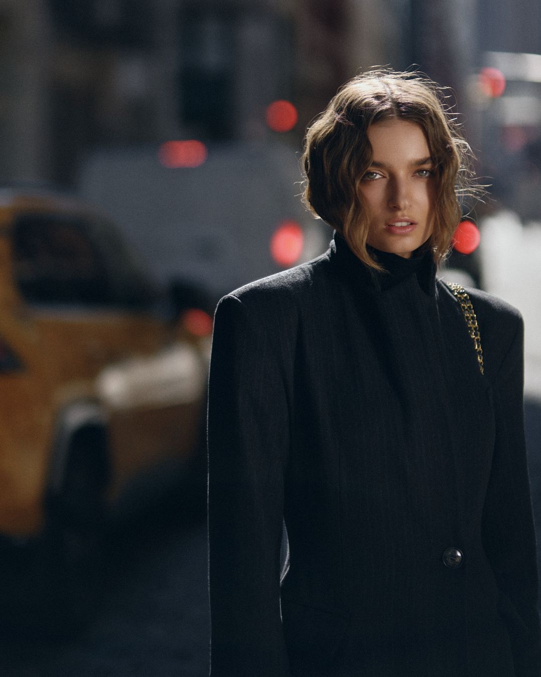 A woman in a black blazer and turtleneck stands on a city street with a yellow taxi in the background.