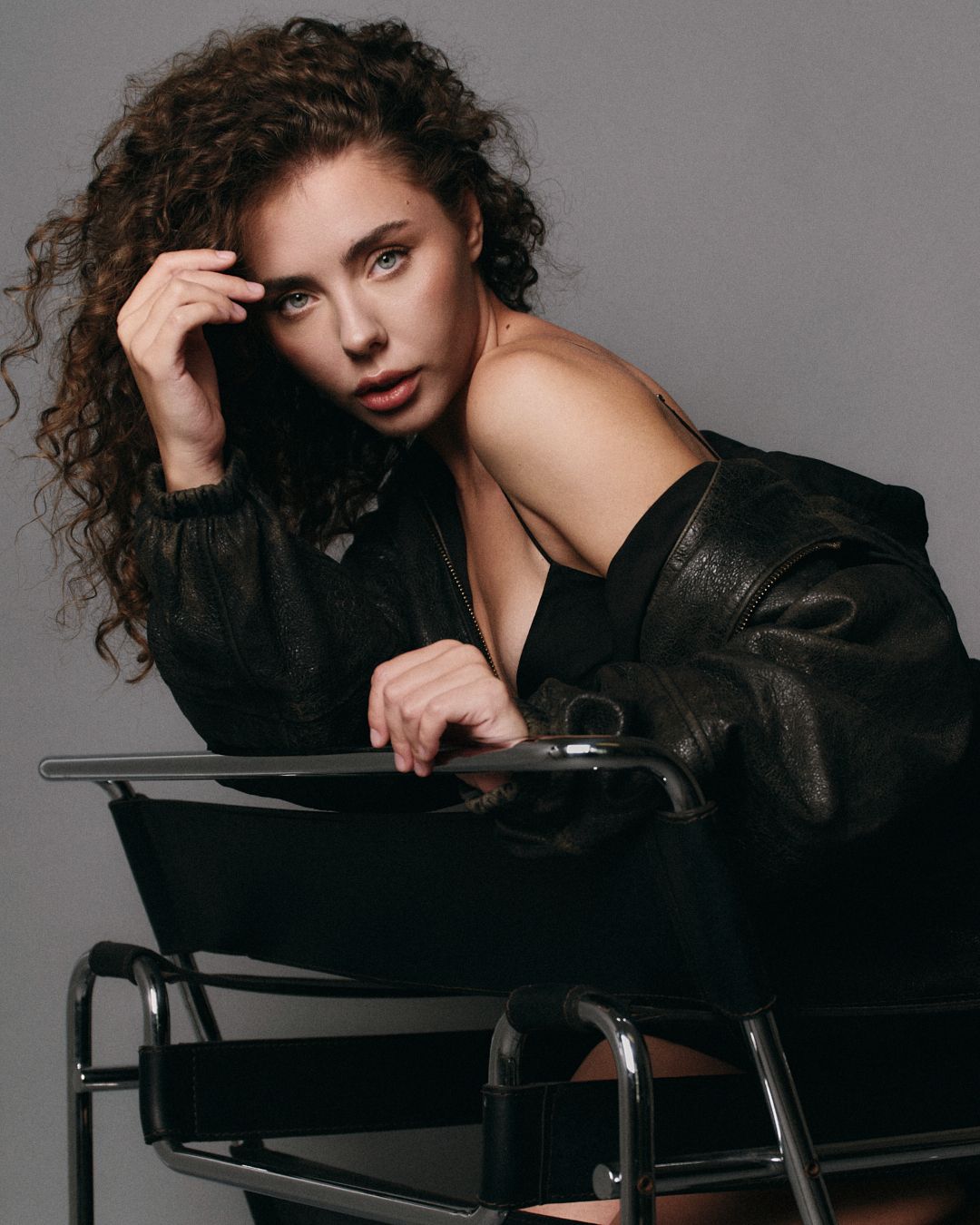 Woman with curly hair in black leather jacket poses on modern chair against gray background.