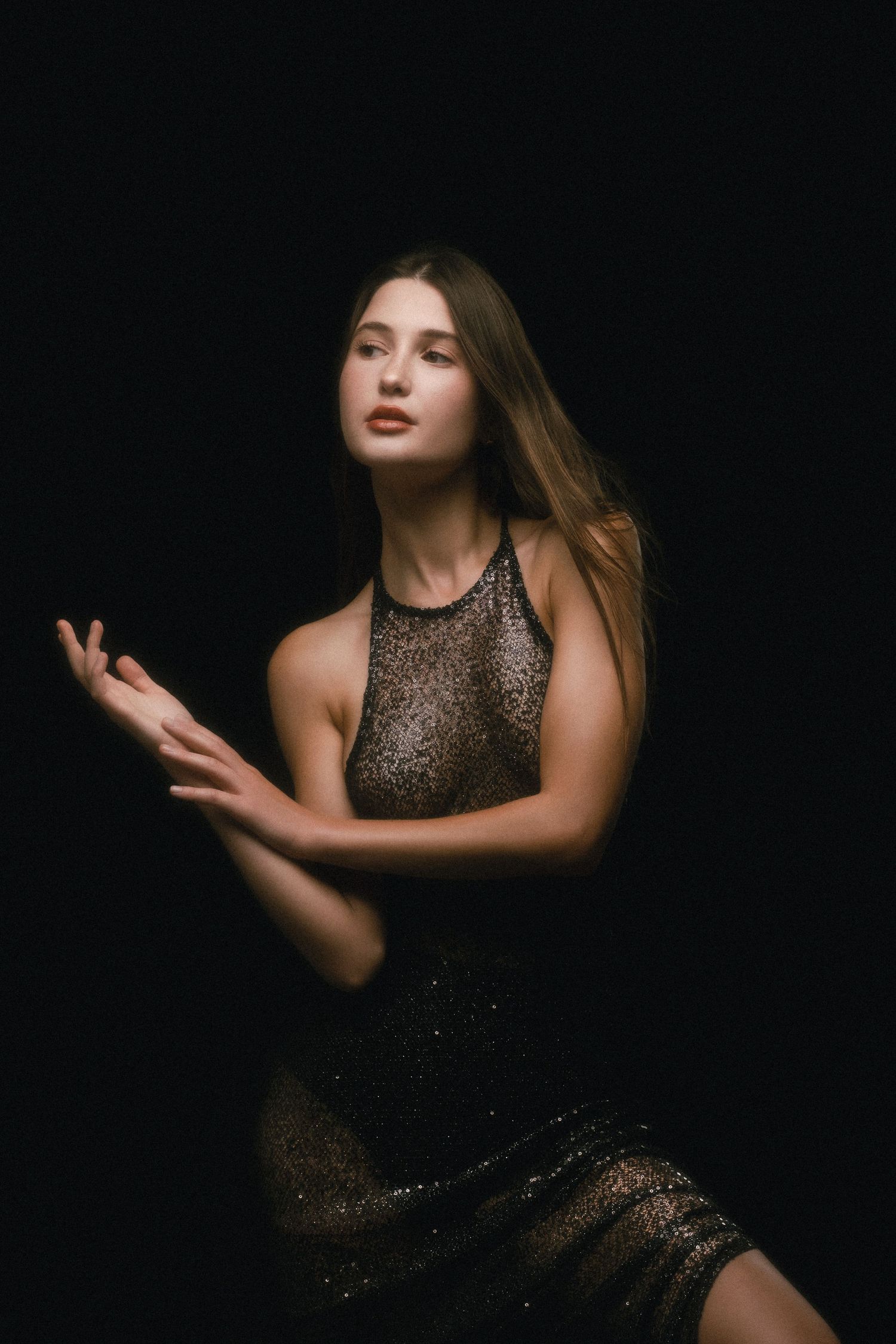 Young woman in a shimmering dark lace dress poses gracefully against a black background in elegant studio portrait.