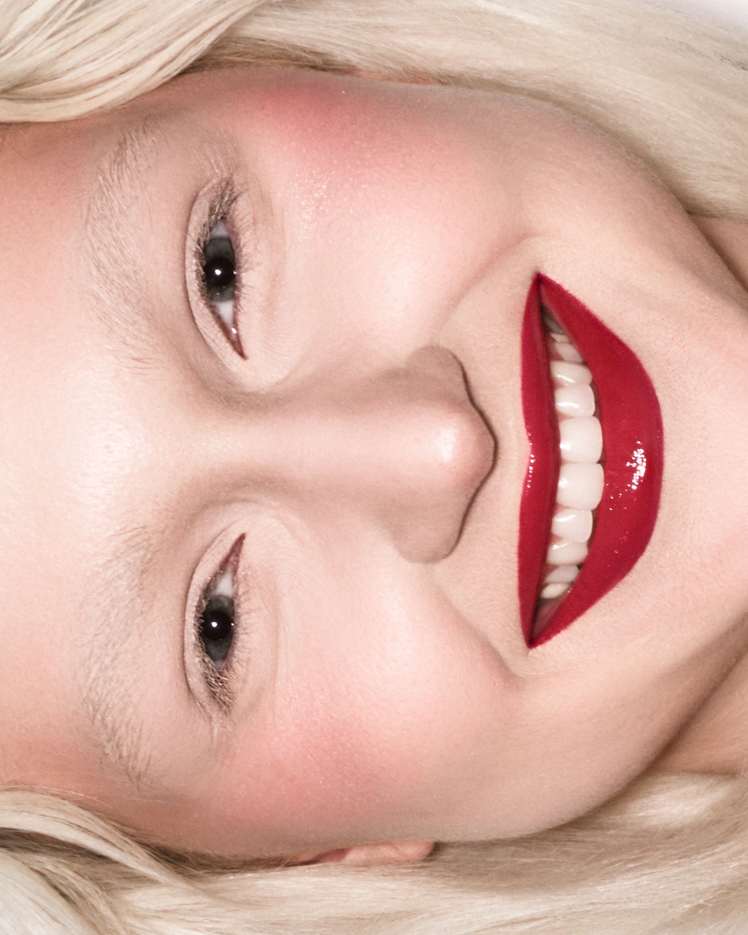 Close-up of smiling blonde woman wearing bold glossy red lipstick and rosy blush makeup.