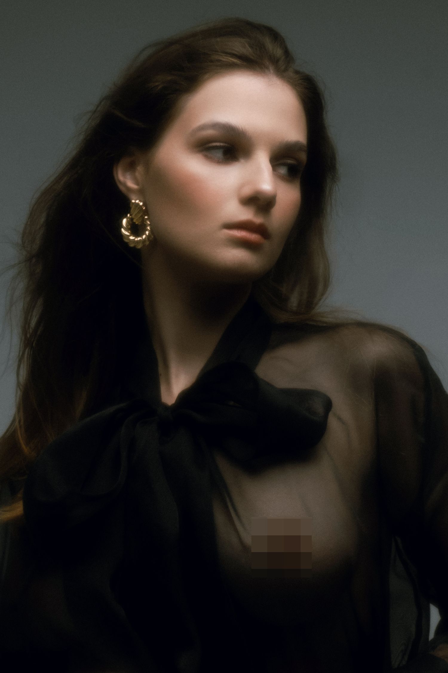 Young woman in sheer black bow blouse with gold hoop earrings, elegant studio portrait photography.