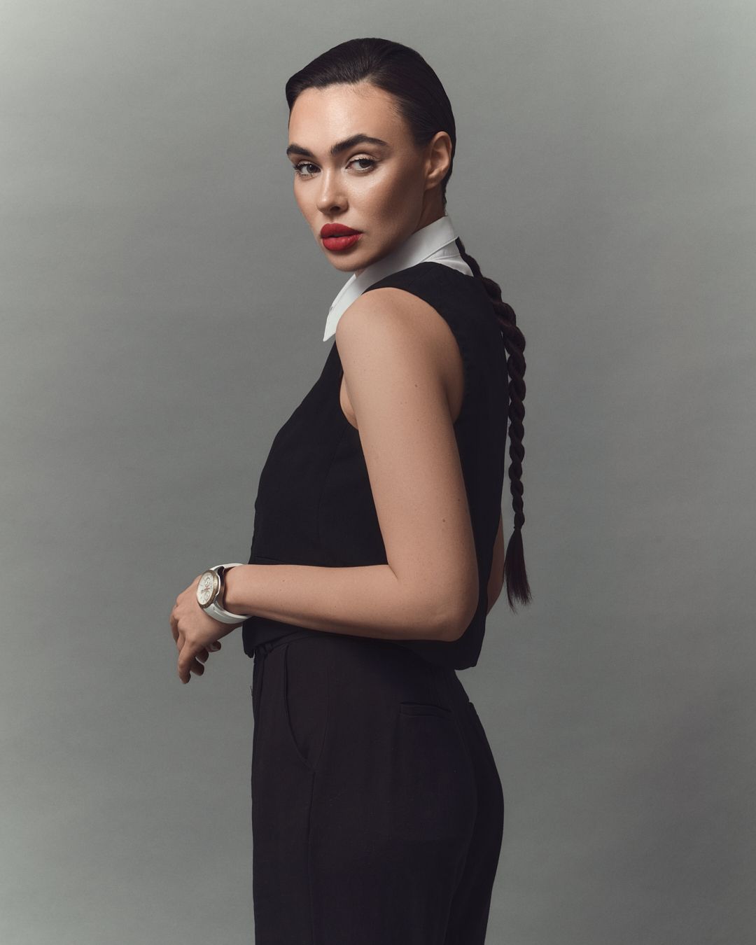 Woman in black sleeveless dress with white collar, red lips, braided hair, and silver watch poses against gray background.