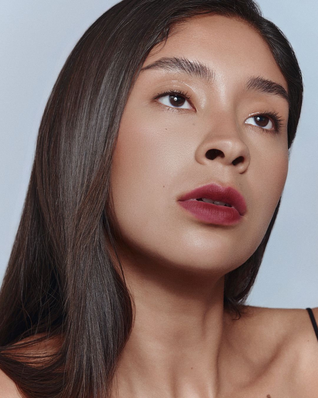 Close-up portrait of a woman with dark hair and bold burgundy lipstick on a light blue background.