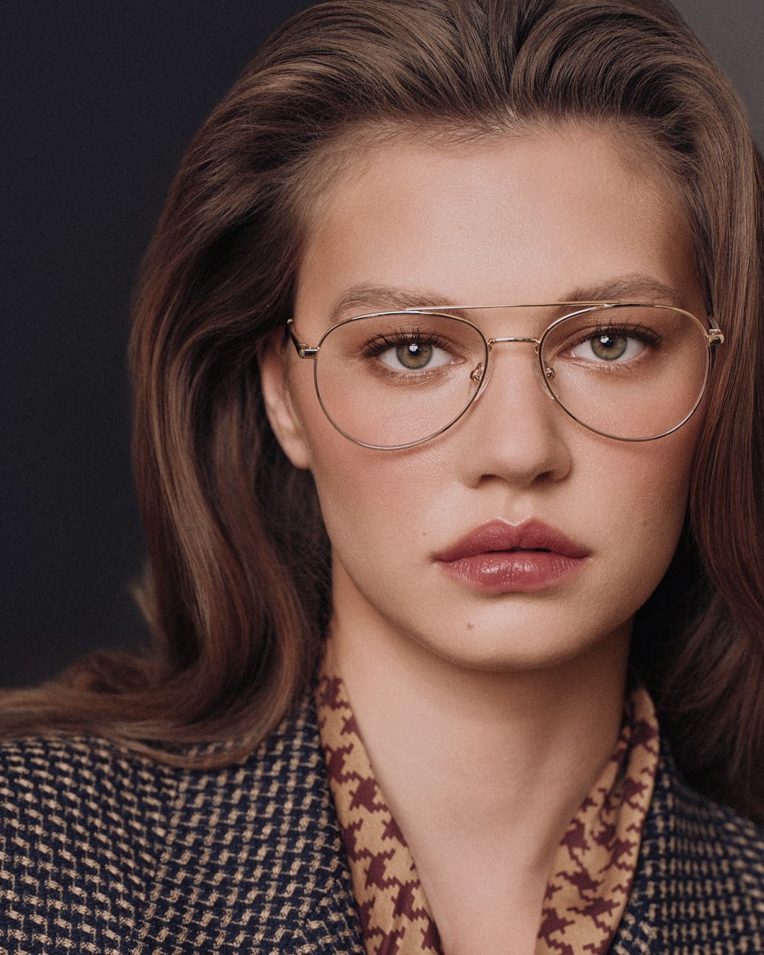 Young woman with brown hair wearing gold aviator glasses and a houndstooth blazer, posing against a dark background.