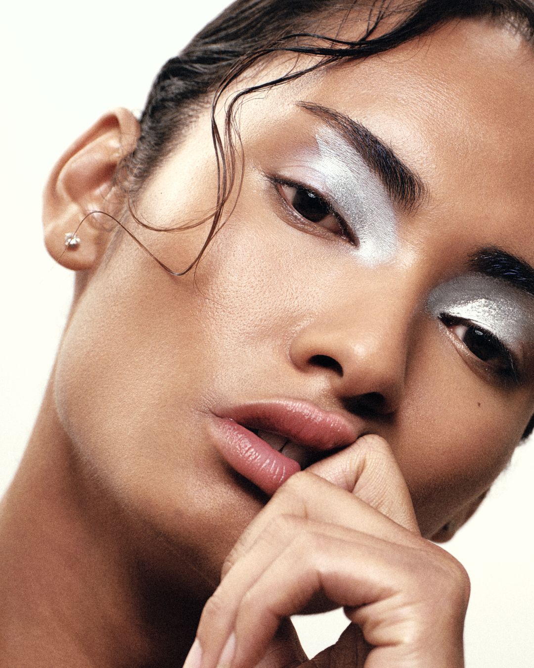 Model with silver metallic eye makeup and wet hair posing with hand near face on light background.