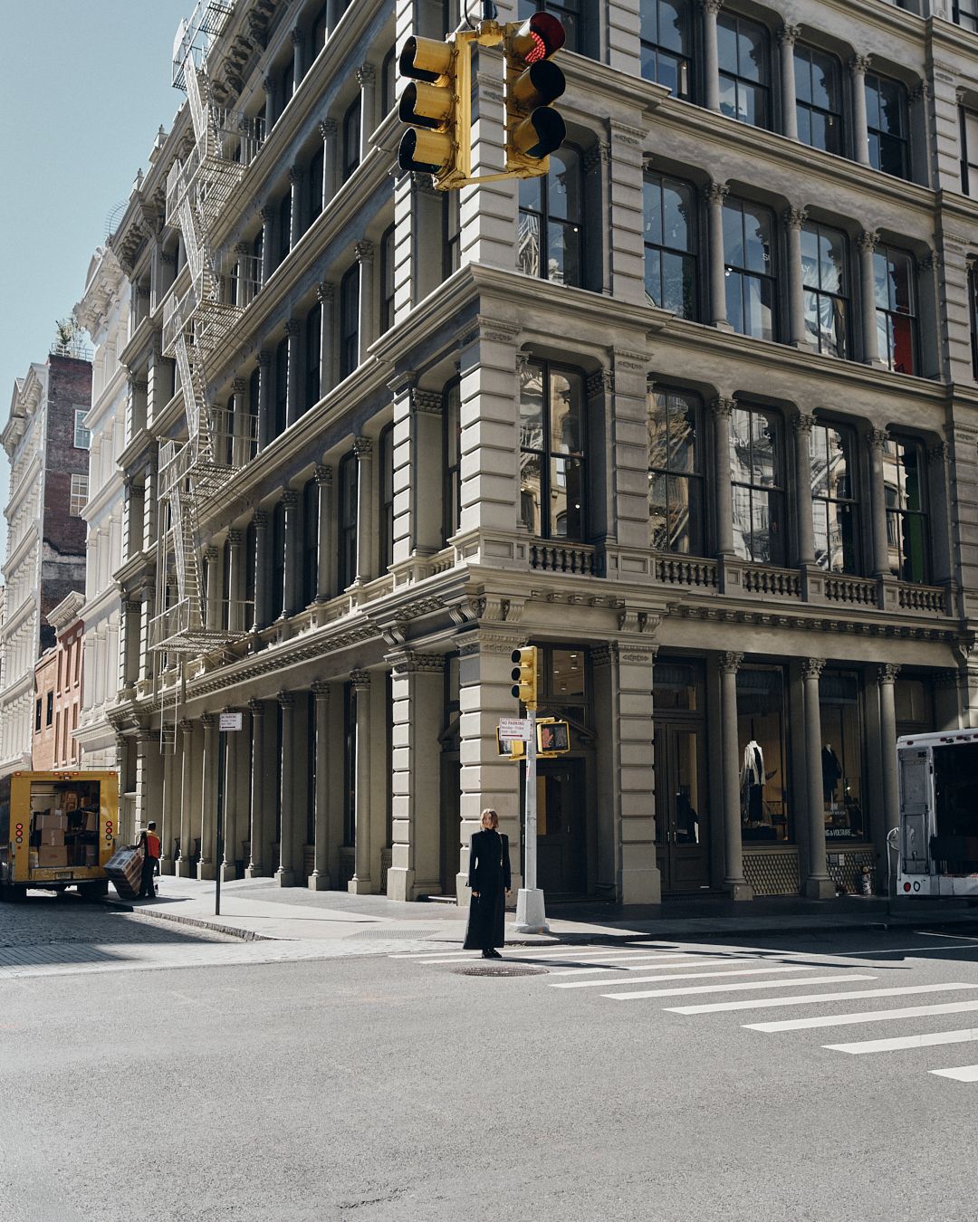 Person standing at a crosswalk in SoHo, NYC, beside a grand beige cast-iron building with large windows and ornate details.