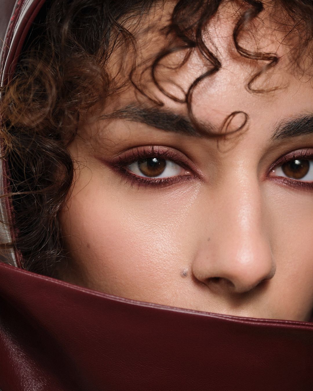 Close-up of a model with curly hair and burgundy smoky eye makeup partially covered by leather hood.