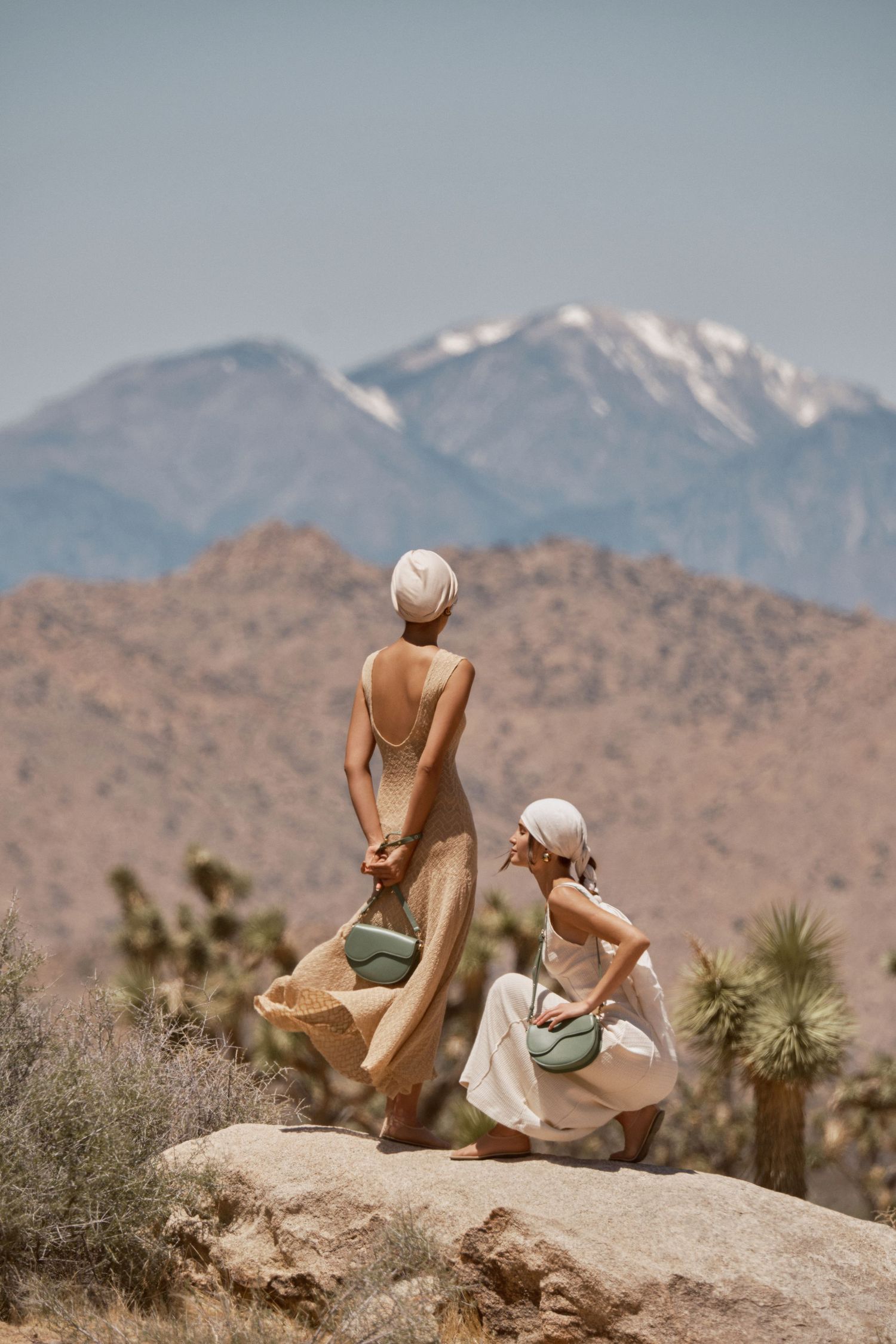 Two women in neutral dresses and head wraps stand on desert rocks with snow-capped mountains in the background.