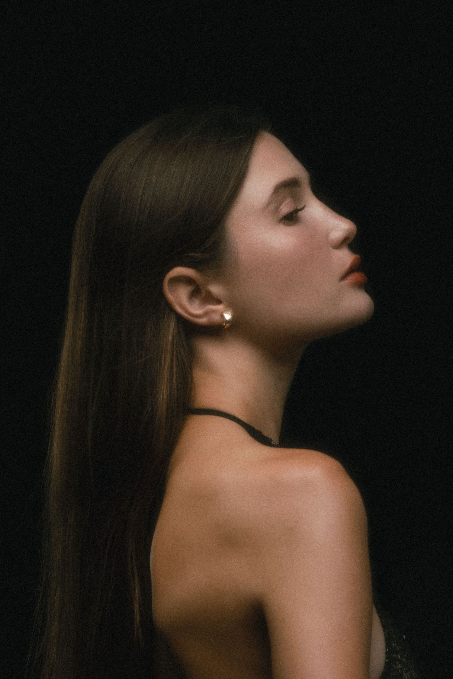 Side profile of a young woman with long straight hair, gold earring, bare shoulders, against a dark background.