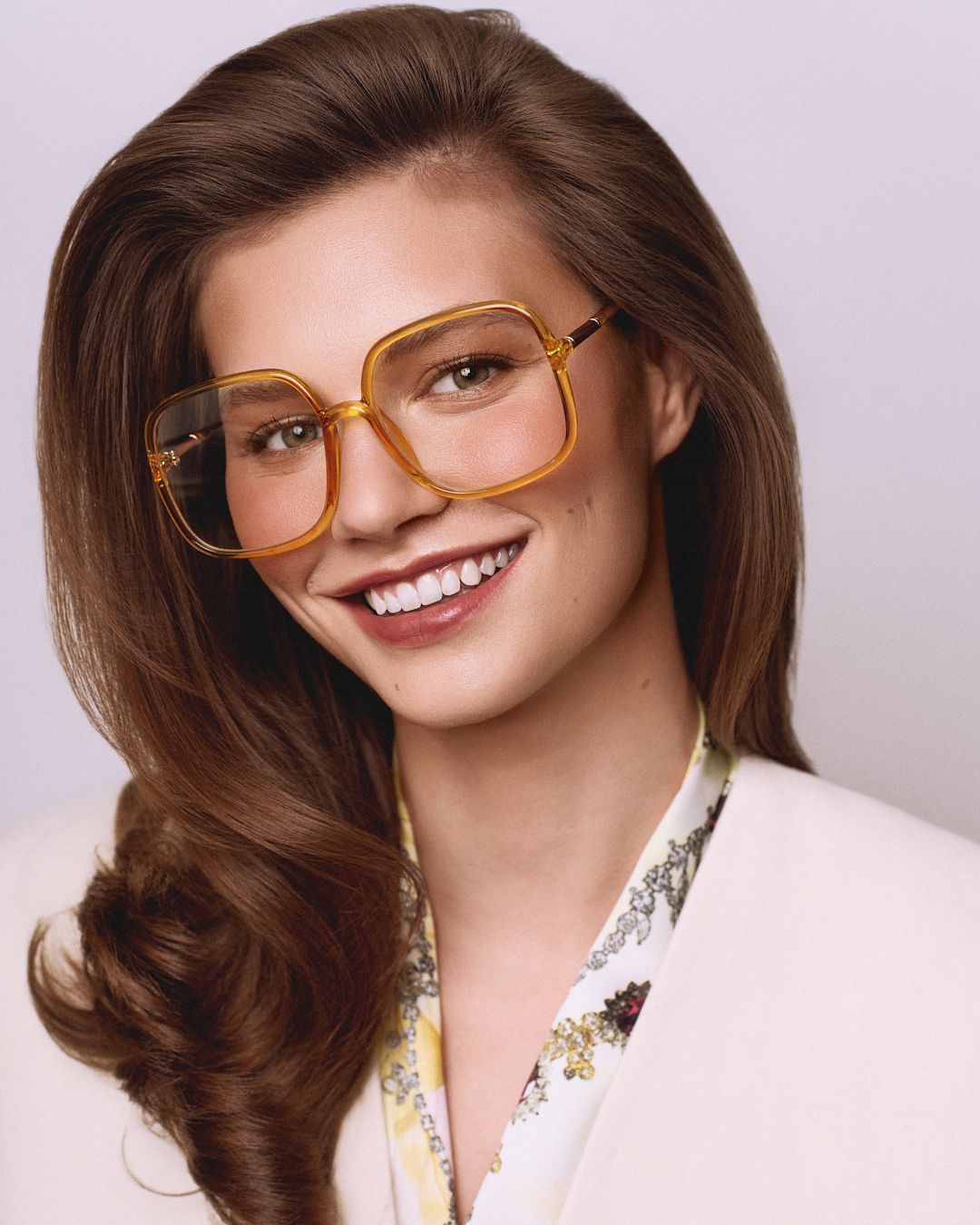 Woman smiling in retro amber square oversized eyeglasses, brown wavy hair, white floral blouse.
