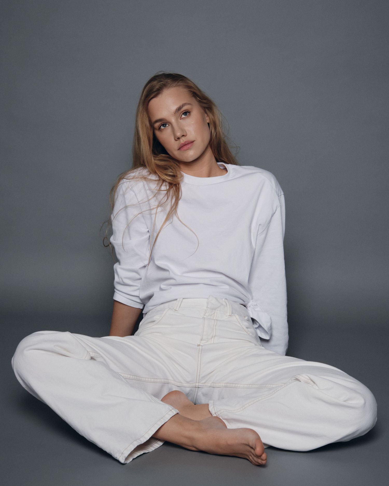 Young woman in white oversized sweatshirt and wide-leg pants sitting cross-legged on floor against gray background.