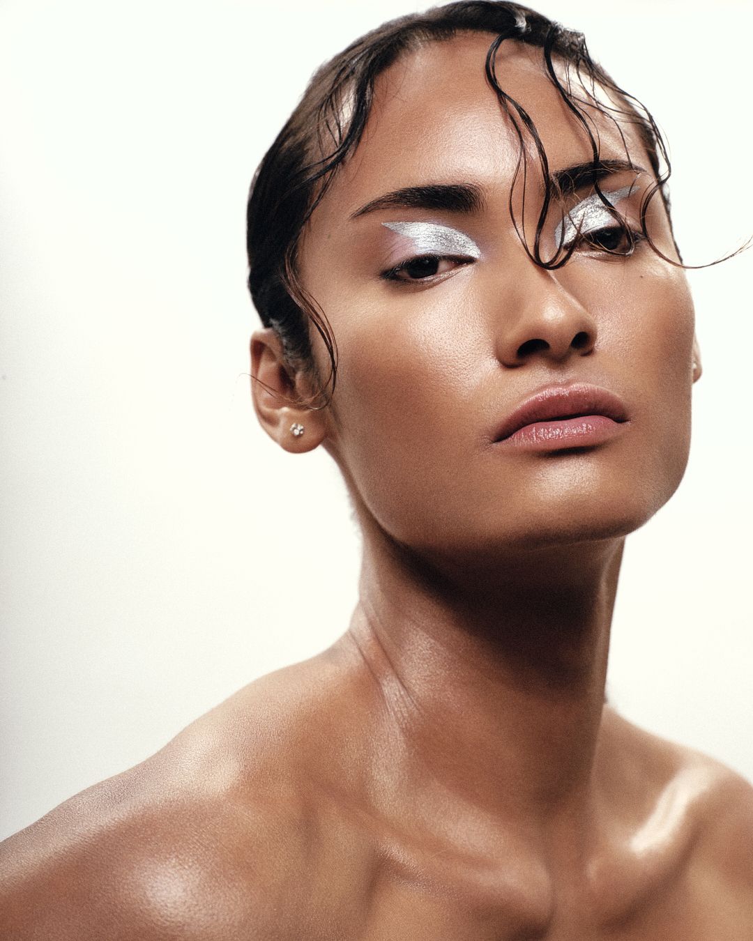 Model with silver metallic eye makeup and wet hair posing against white background.