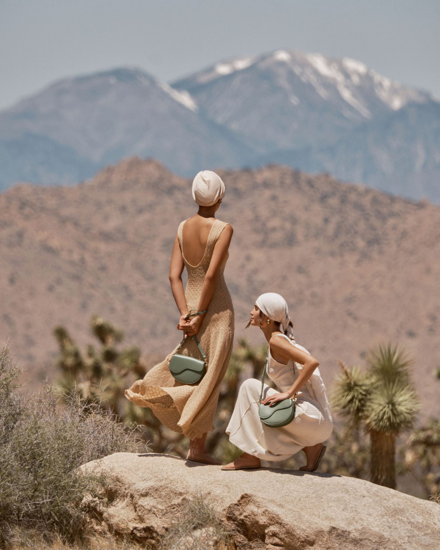 Two women in neutral dresses holding bags on desert rocks with snow-capped mountains backdrop.