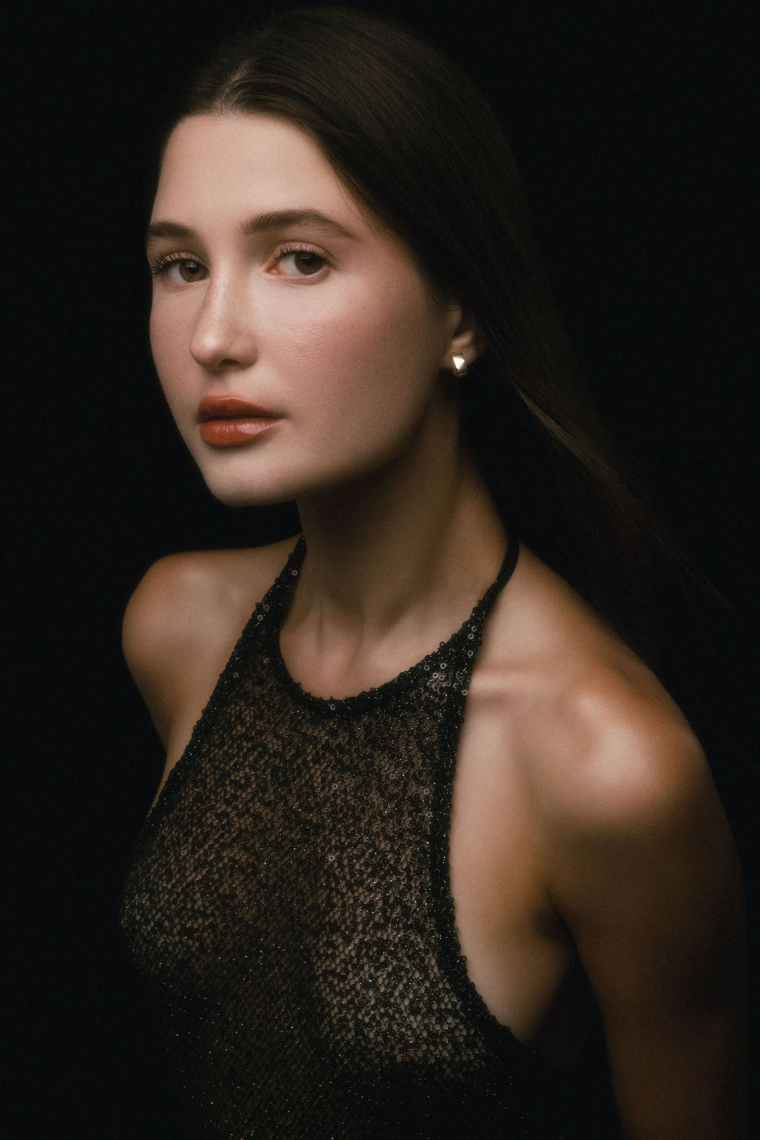 Young woman with dark hair wearing a black sequin halter top, pearl earring, posing against dark background.