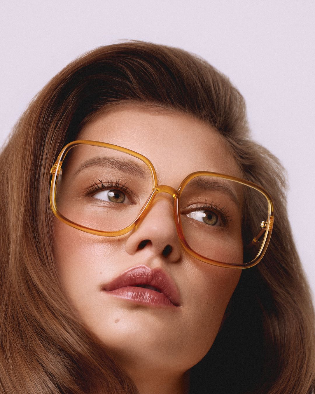 Young woman wearing oversized amber square eyeglasses, looking up against a soft lavender background.