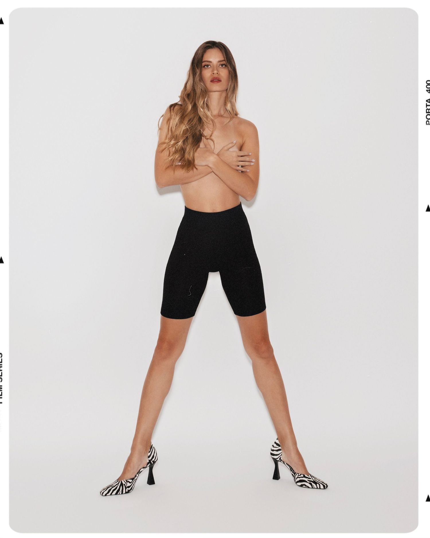 Model wearing black cycling shorts and zebra print heels, posing with arms crossed over chest against white background.