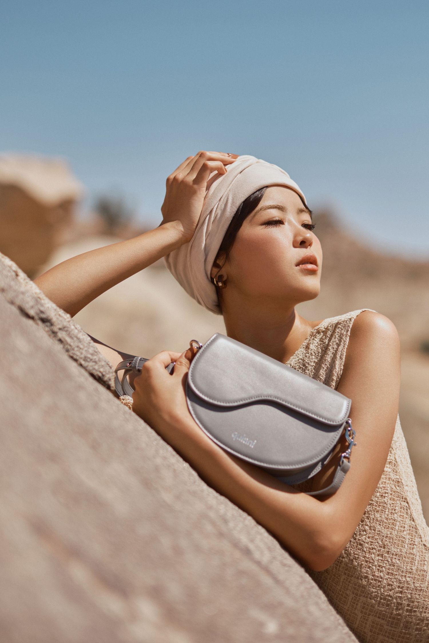 Woman in beige turban holding a gray suede crossbody bag, posing against desert rocks under a clear blue sky.