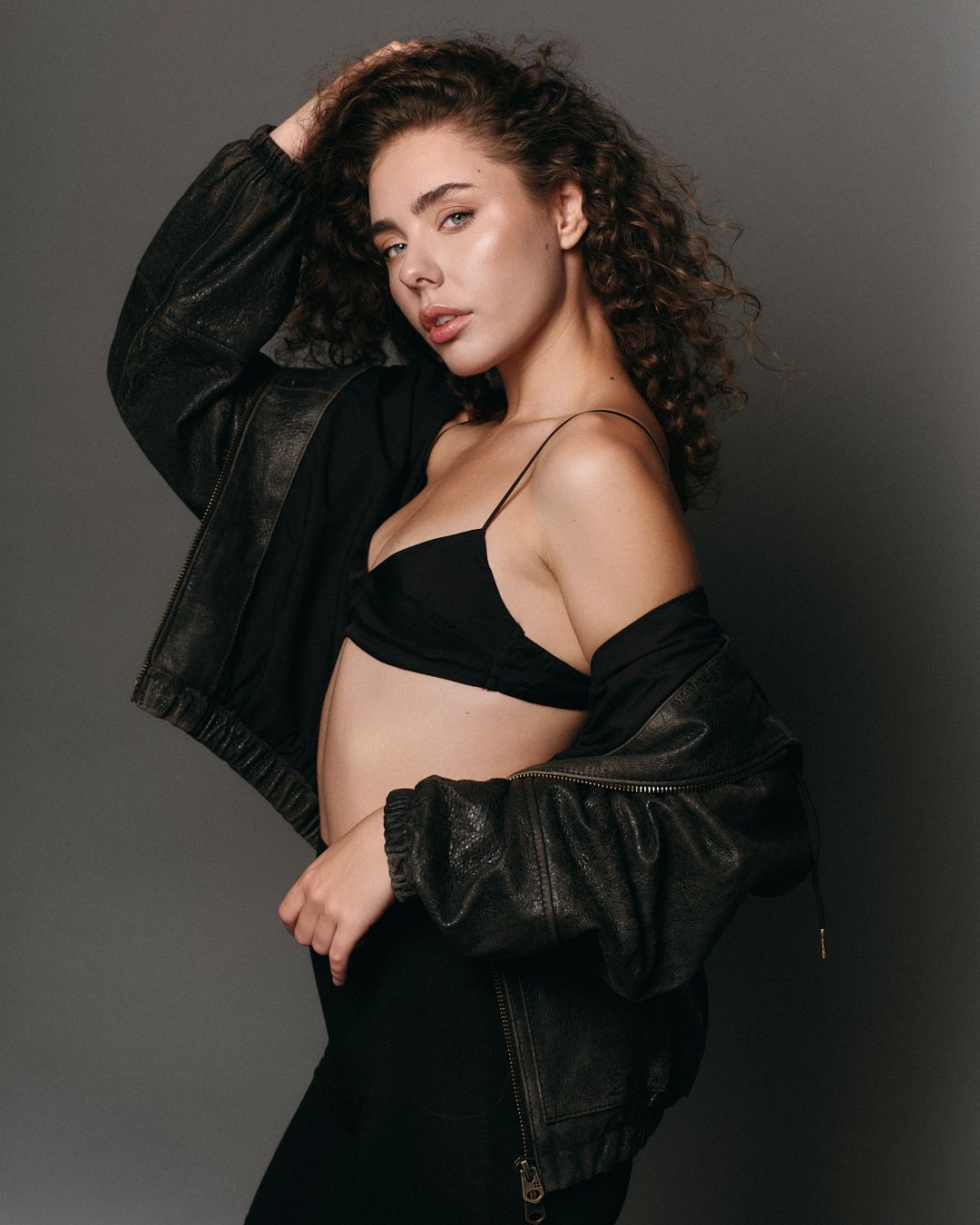 Young woman with curly hair poses in black bralette, open leather jacket, and dark pants against gray background.