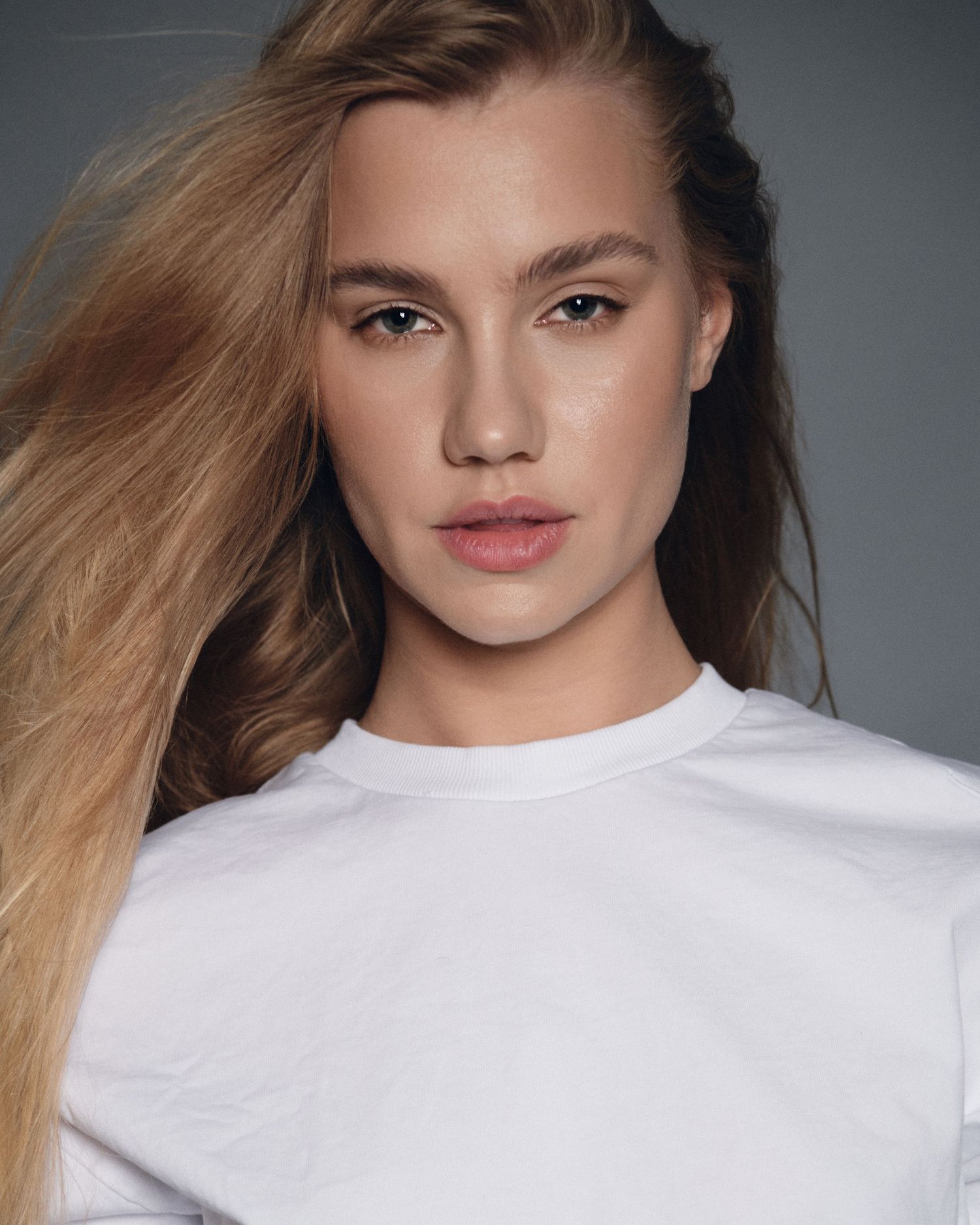 Young woman with long blonde hair wearing a white t-shirt poses against a gray background in a clean beauty shot.