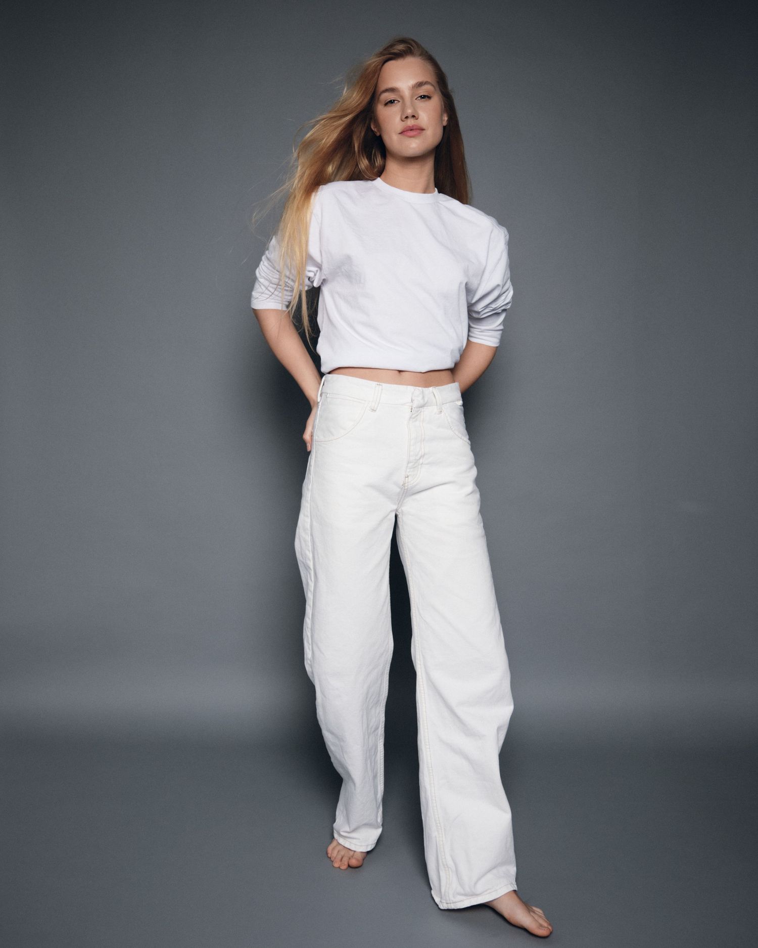 Woman in white crop tee and wide-leg white jeans poses barefoot against gray background.