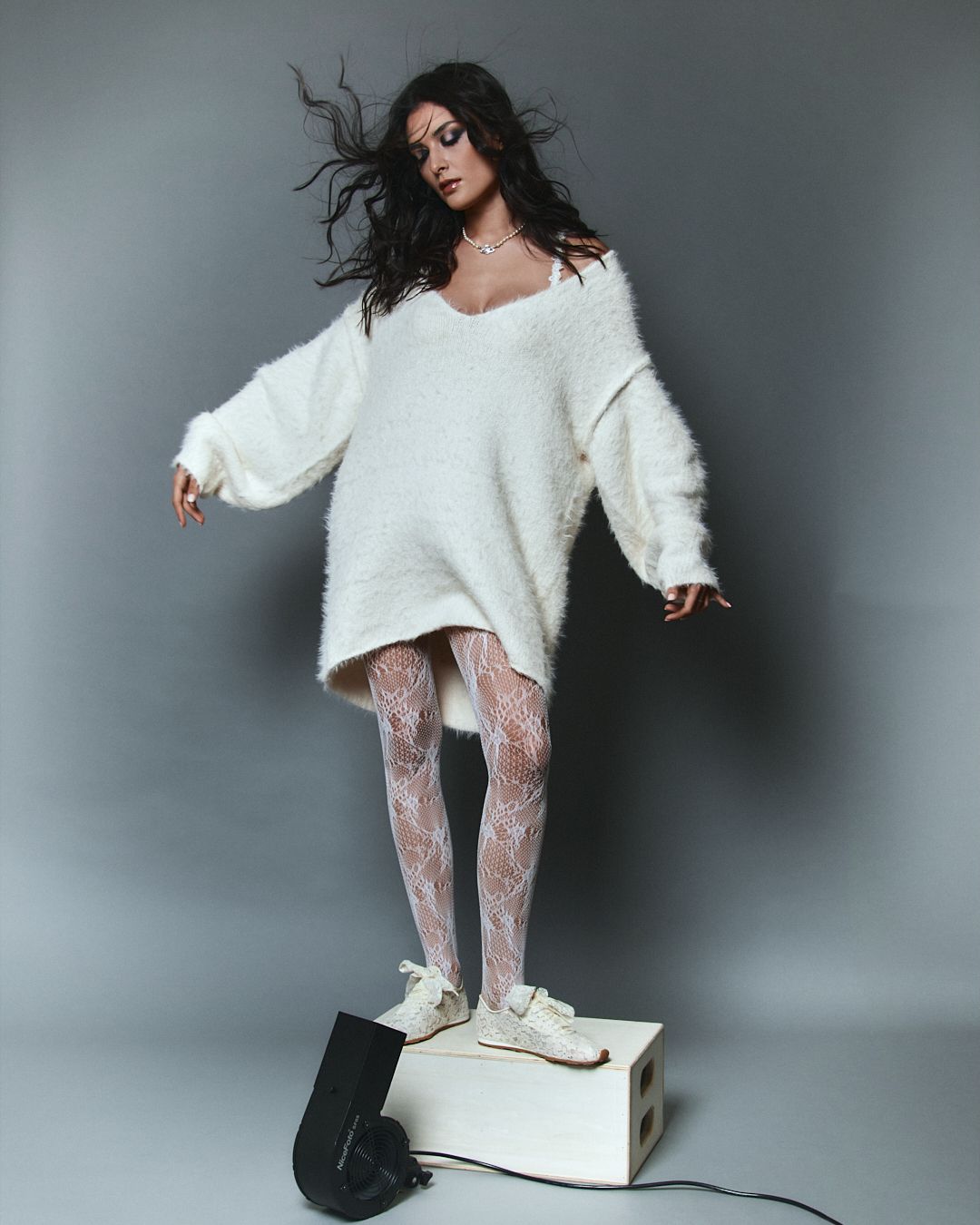 Woman in oversized white sweater dress, lace tights, and white sneakers poses on a white box.
