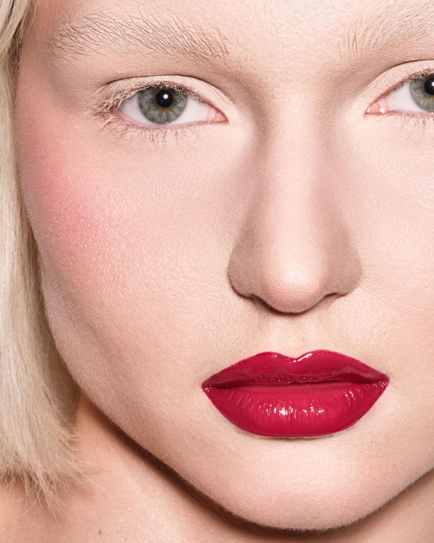 Close-up of blonde model with green eyes wearing vibrant glossy red lipstick and subtle blush.