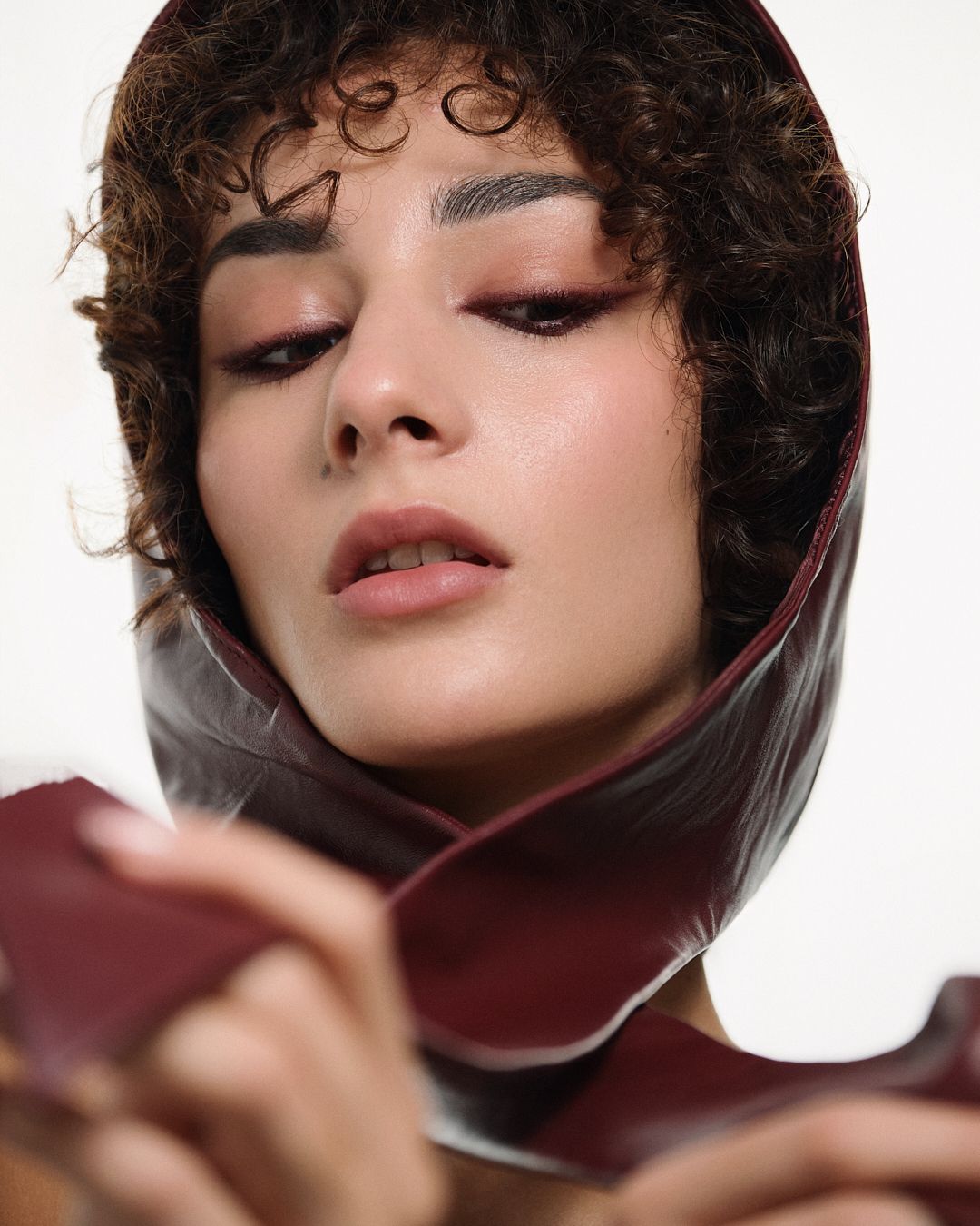 Close-up of a model with curly hair wearing a burgundy leather hood, eyes closed, editorial look.