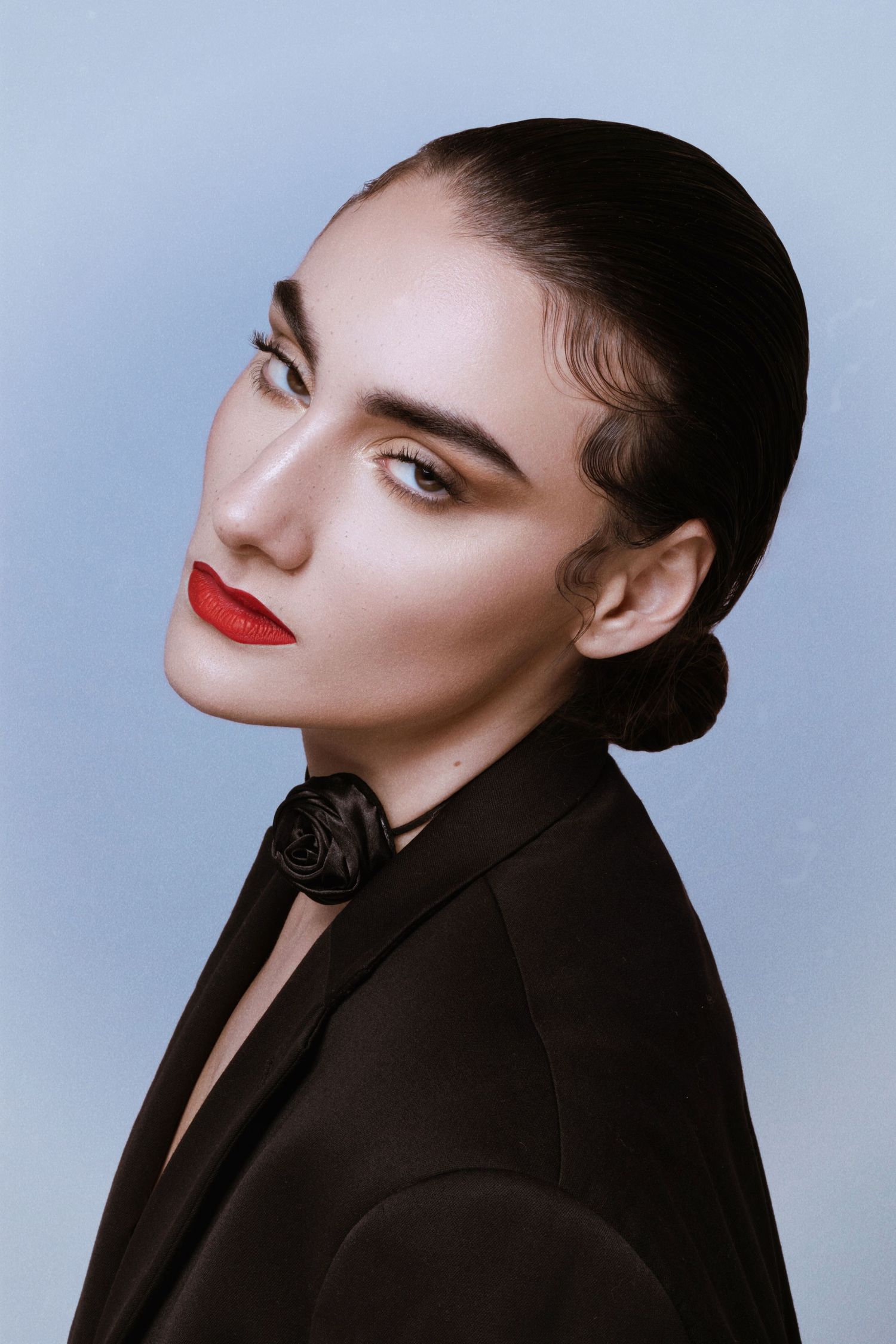 A woman with sleek dark hair in a bun, bold red lips, and dramatic brows wearing a black blazer with a rose detail.