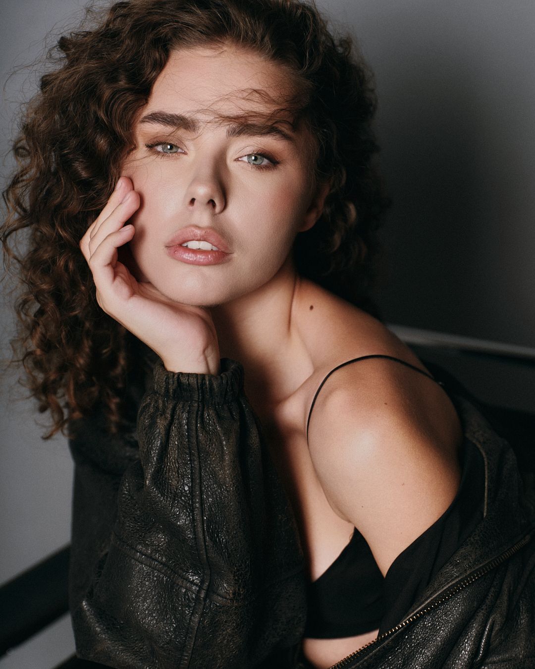 Young woman with curly brown hair and blue eyes poses in black leather jacket for a professional portrait.