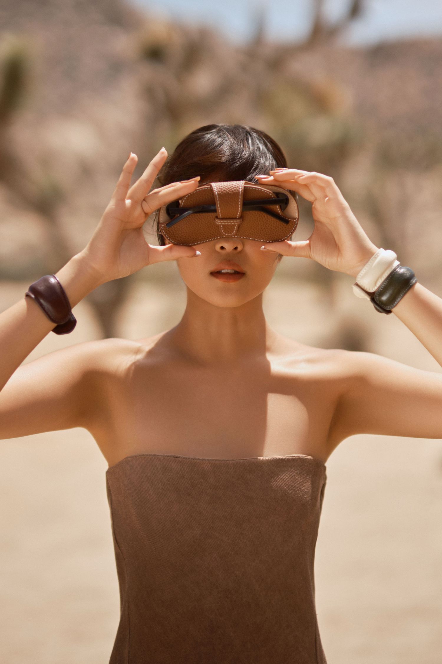 Woman in desert wearing brown VR headset and bangles, holding device to eyes against arid landscape backdrop.