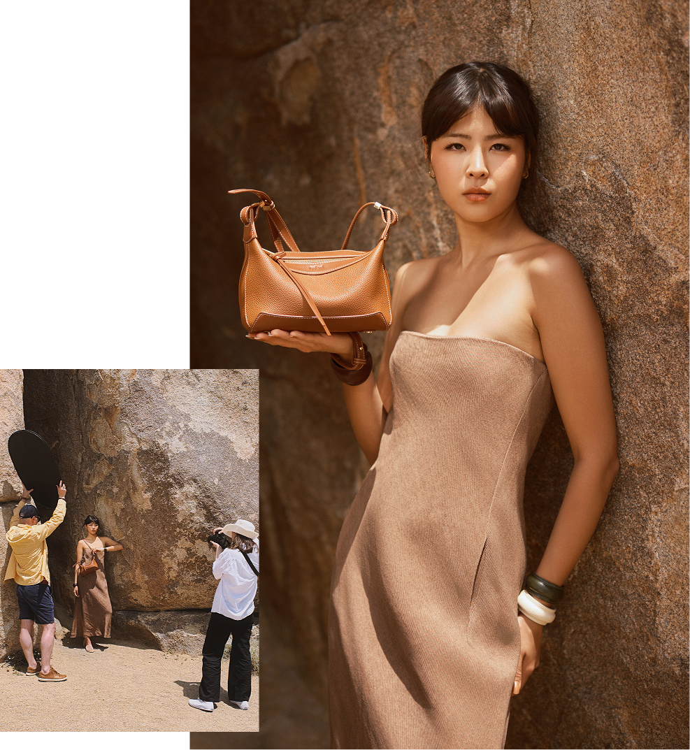 Woman in beige strapless dress holding tan leather shoulder bag against stone wall.