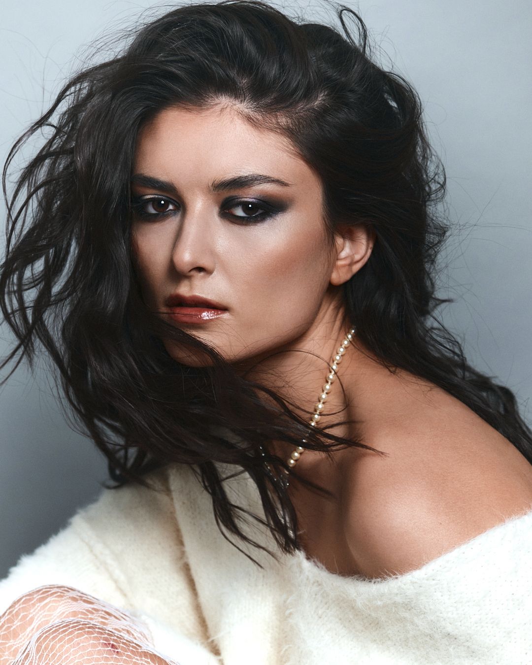 Dark-haired woman in white fur wrap with pearl necklace and smoky eye makeup.