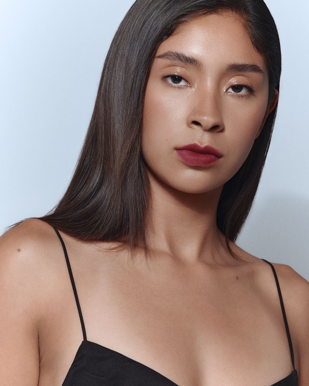 Young woman in a black cami top with dark hair and deep red lipstick against a light background.