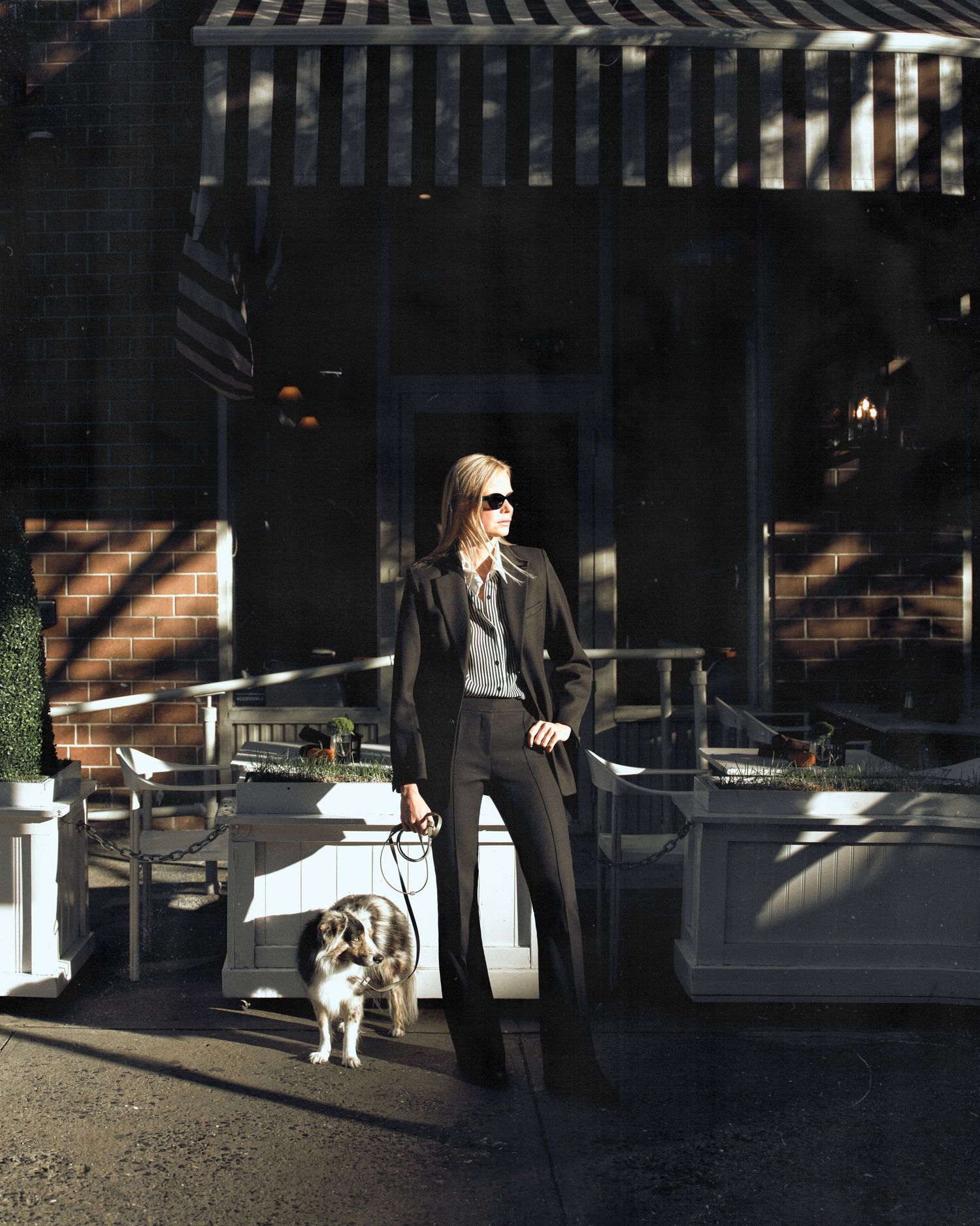 Woman in black suit walks dog on leash outside a restaurant patio in bright sunlight.