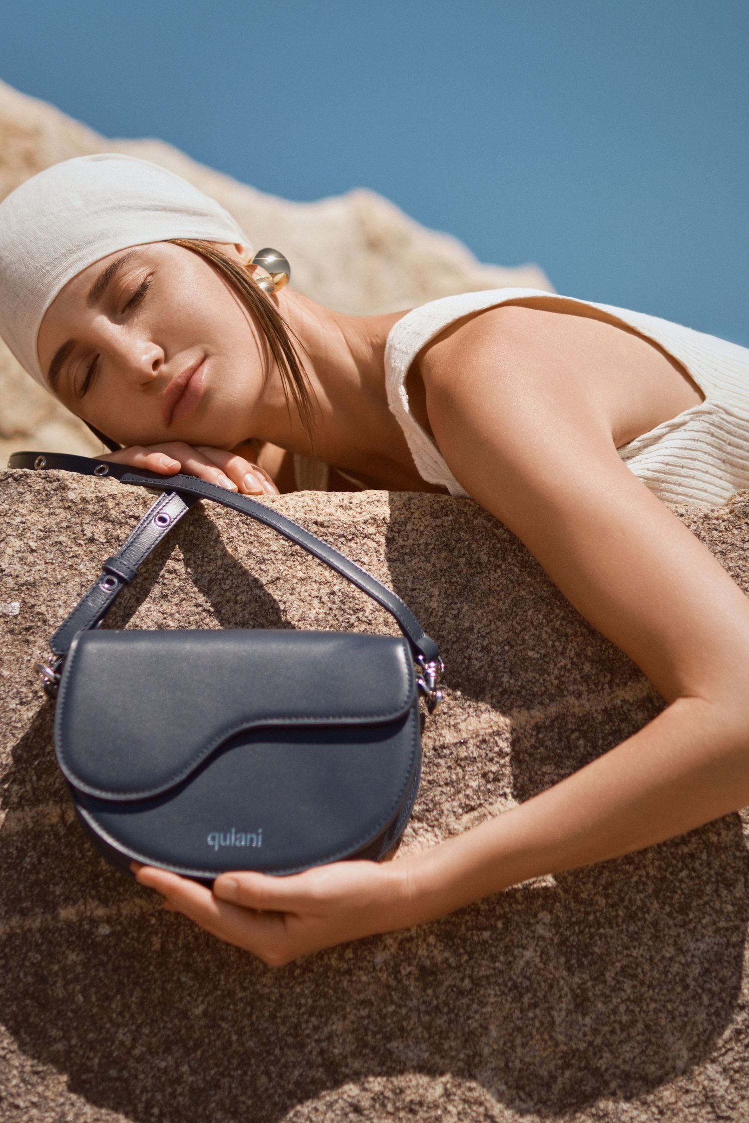 Woman resting on rocks with a navy blue Quilani leather saddle bag in a sunny outdoor setting.