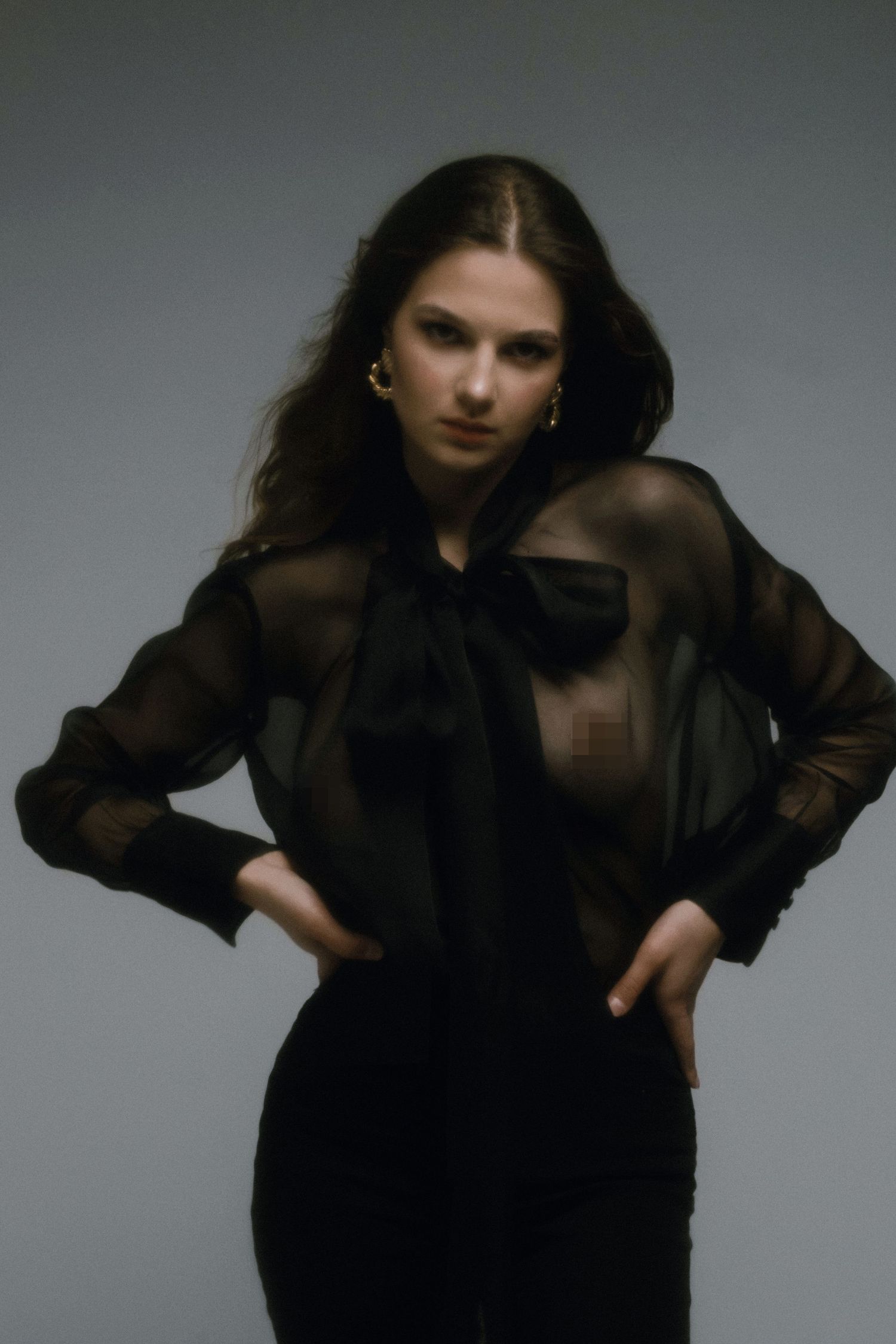 Young woman in sheer black blouse with bow tie and dark skirt poses confidently against gray background.