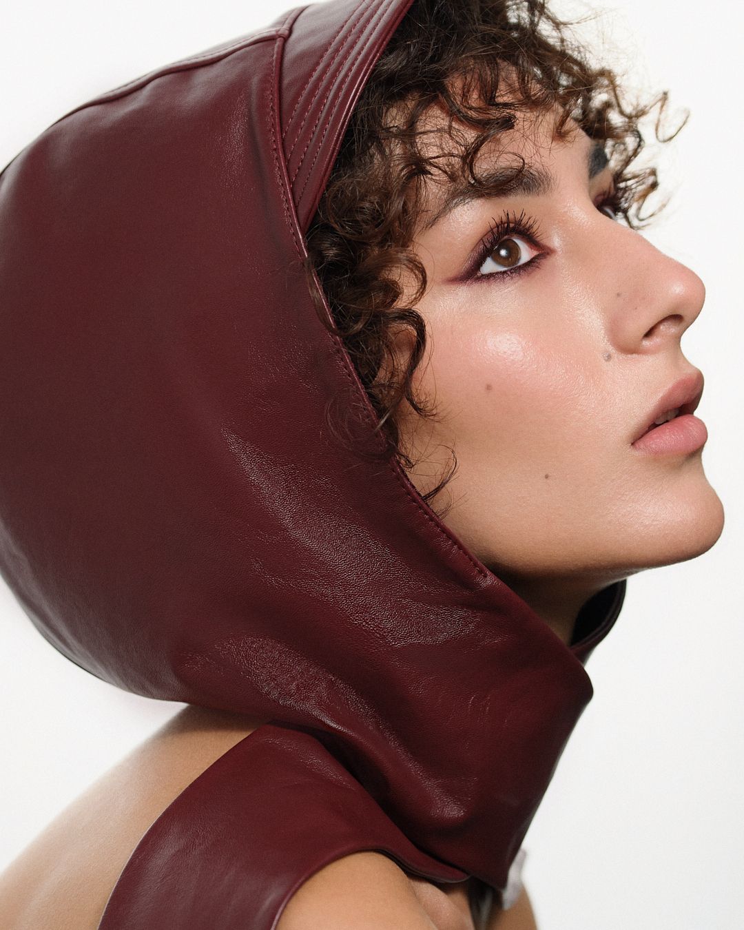 Side profile of a model with curly hair wearing a burgundy leather bonnet in an editorial shoot.