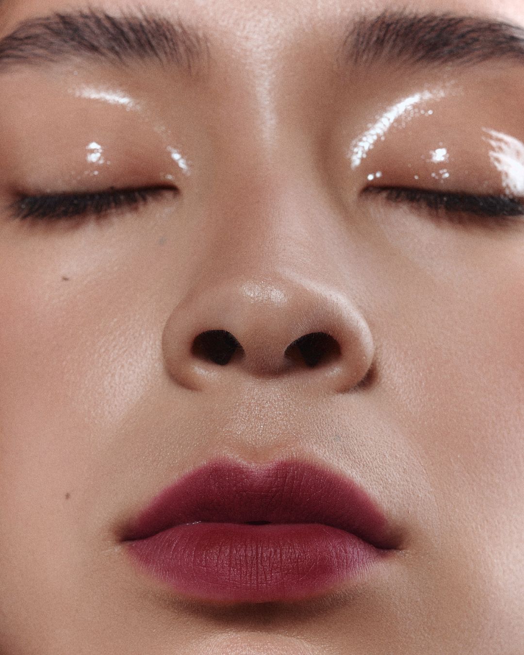 Extreme close-up of a face with glossy eyelids and burgundy lips showcasing a dewy makeup look.