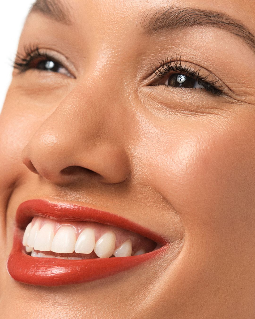Close-up of a woman's bright smile with bold red lipstick, glowing skin, and long lashes.
