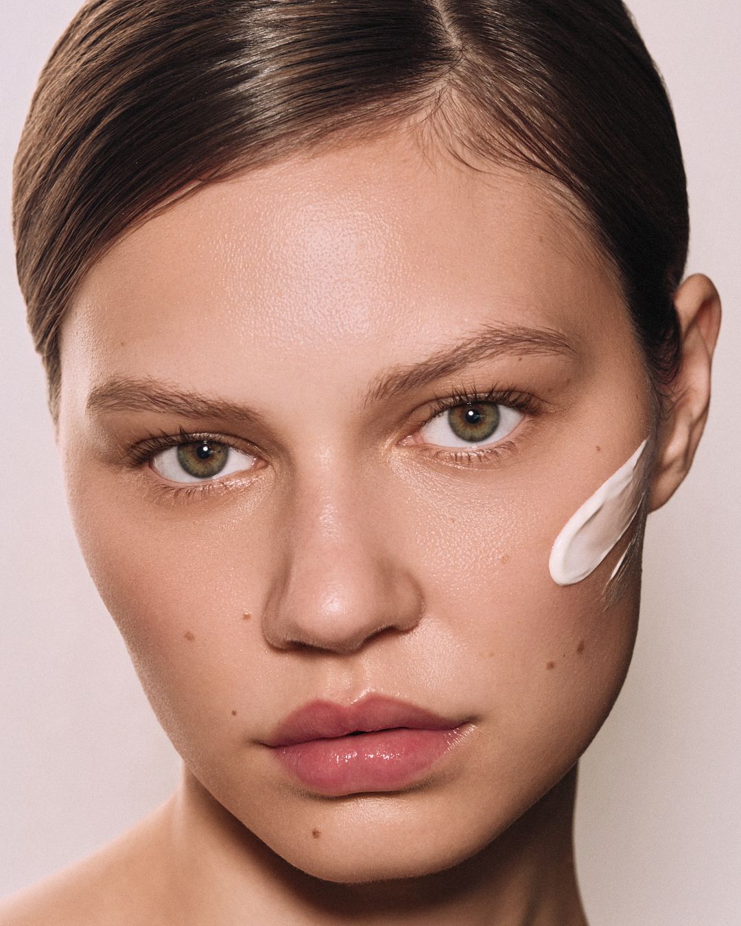 A woman with green eyes and pulled-back hair with a white cream swatch on her cheek against a neutral background.