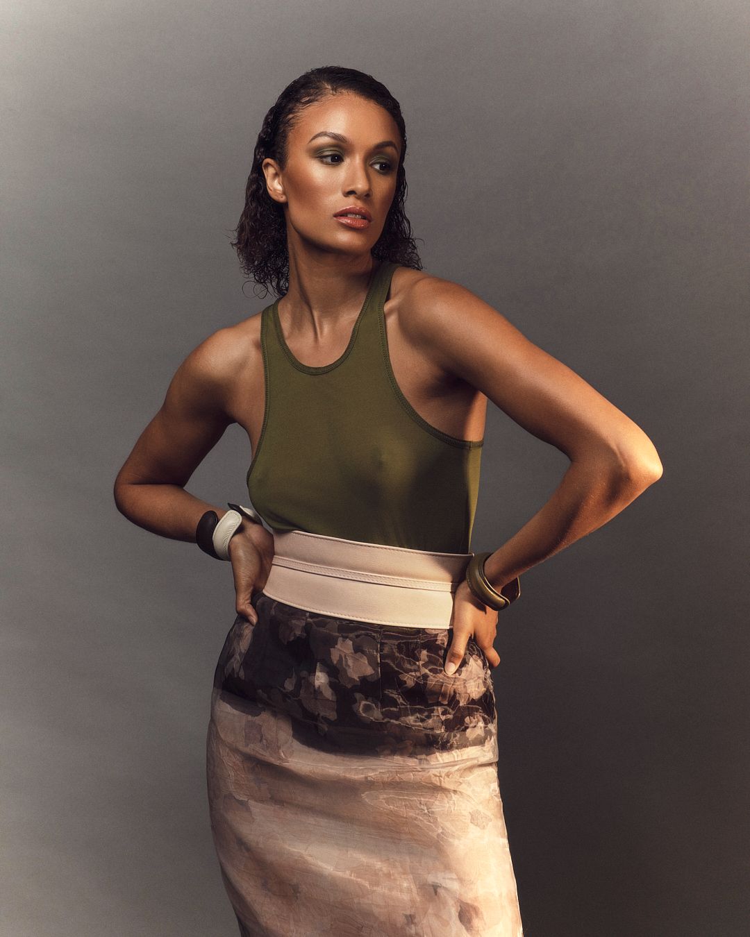 Woman in olive green tank top and abstract print skirt with white belt poses confidently against gray background.