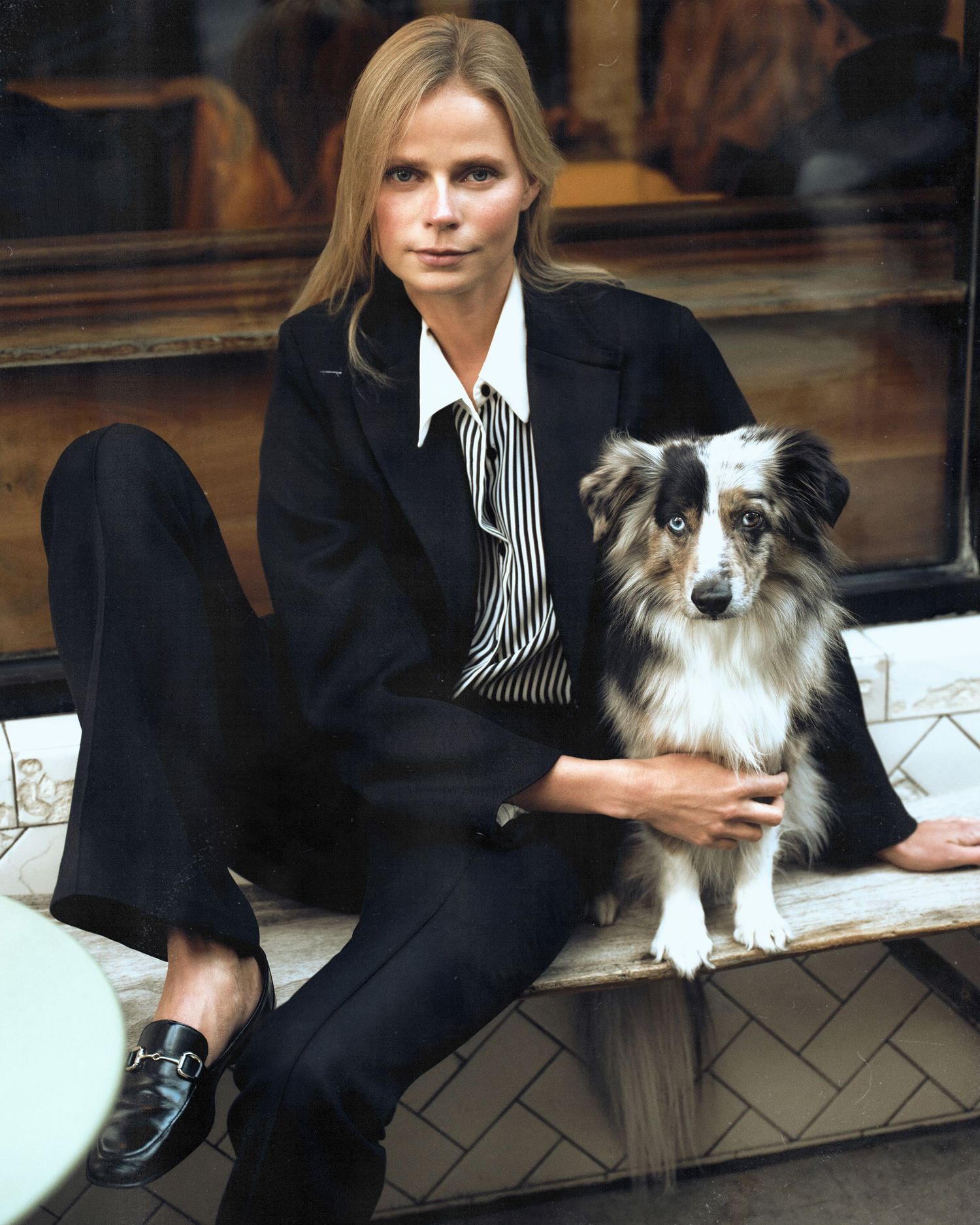 Woman in black blazer sitting with an Australian Shepherd dog on a bench, wearing loafers and striped shirt.