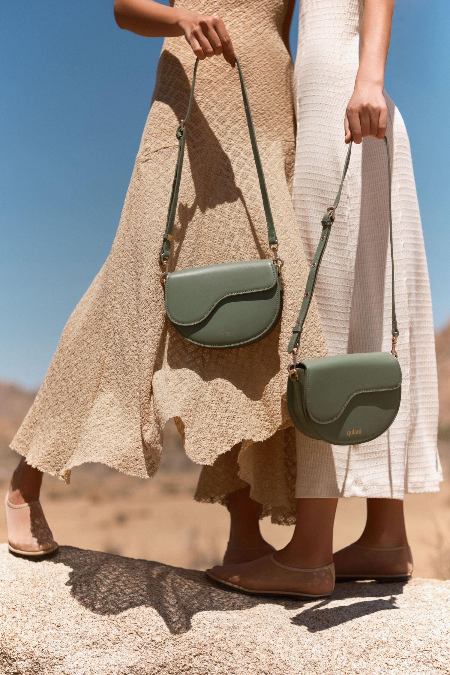 Two women in beige dresses holding sage green crossbody saddle bags outdoors in a desert setting.