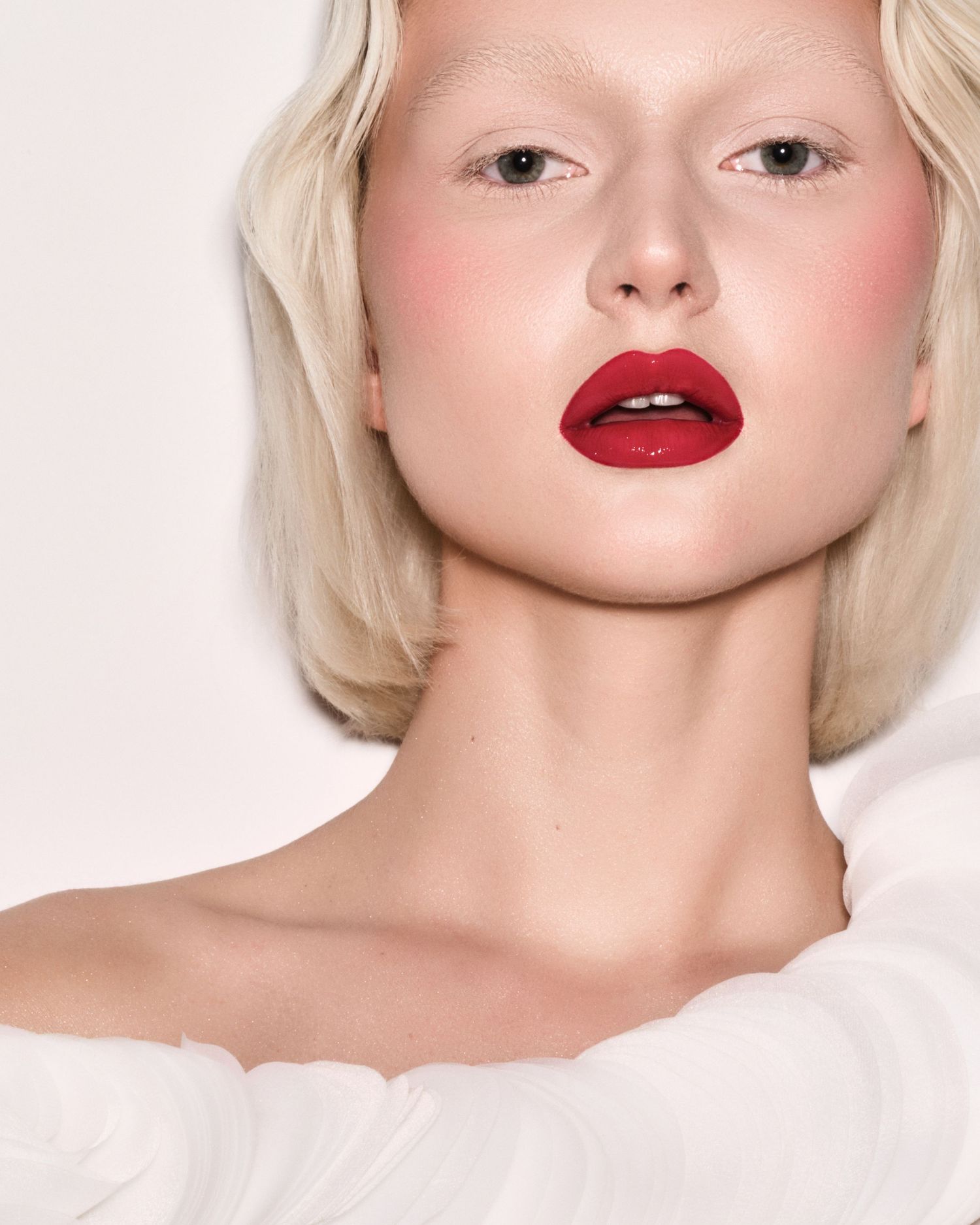 Blonde model in white top wearing bold red glossy lips and rosy blush against light background.